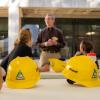 Dean Keith Molenaar stands before a group of people sitting at tables outdoors, with yellow construction hats on the tables.